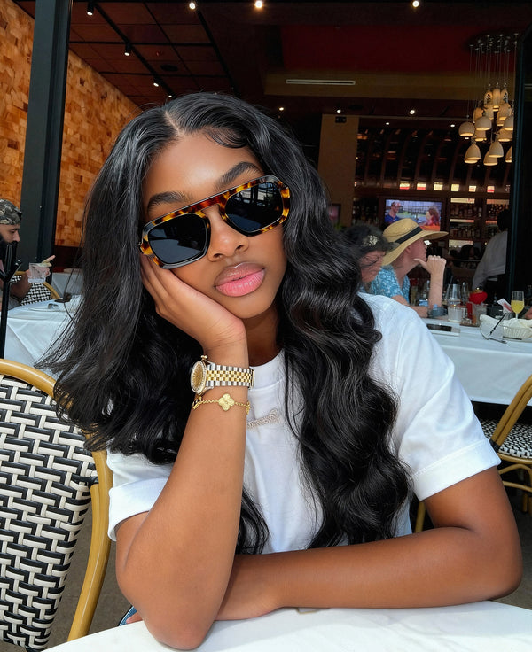 Woman wearing sunglasses and a white shirt in a restaurant setting wearing raw indian hair bundles